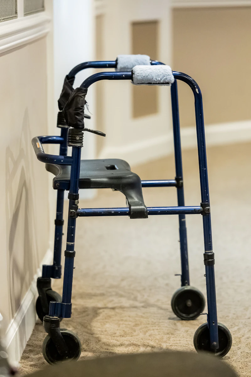 A blue folding walker with a seat and wheels standing in a carpeted hallway